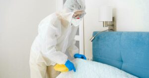 A person in a white biohazard suit with goggles and a mask, lifting and cleaning a mattress