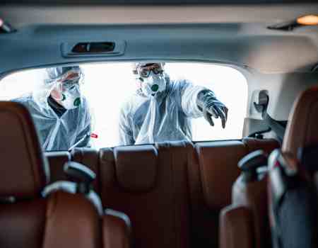 A view of workers from the inside of a vehicle. They wear protective gear and are about to disinfect the back seats.