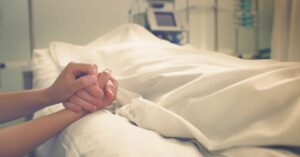 A person holding the hand of a deceased individual in a hospital bed that has a sheet placed over them.