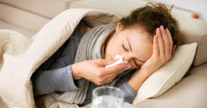 A woman lying on a couch and blowing her nose. She is wrapped in covers and has a cup of water next to her.