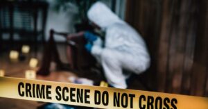 A crime scene cleaner wearing biohazard gear crouches down. Crime scene tape reading "CRIME SCENE DO NOT CROSS" blocks the room.