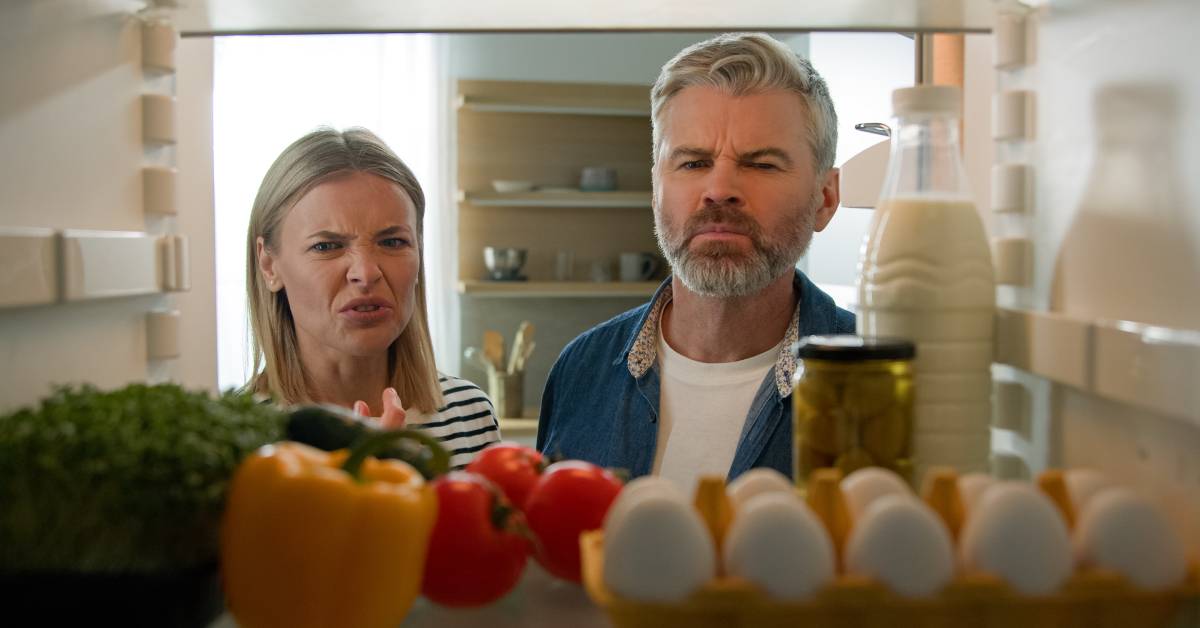 Two people looking inside of a refrigerator with a look of disgust. There are vegetables, milk, and eggs inside.