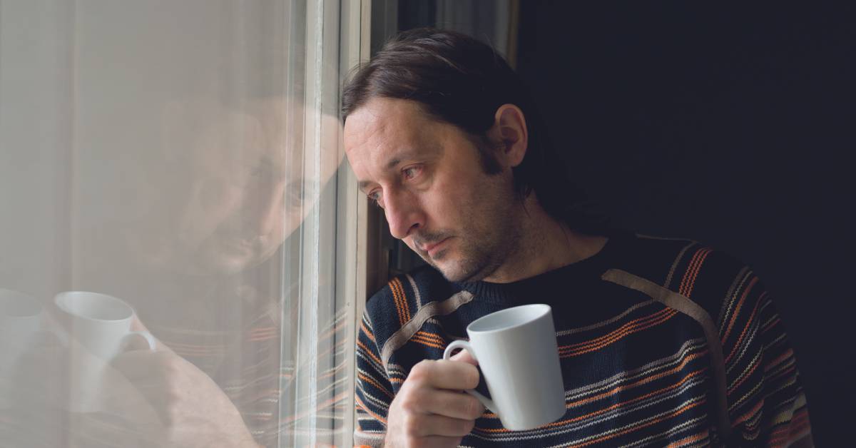 A man leaning on a window looking sad and holding a glass mug. He is in a dark room and wearing a stripped sweater.