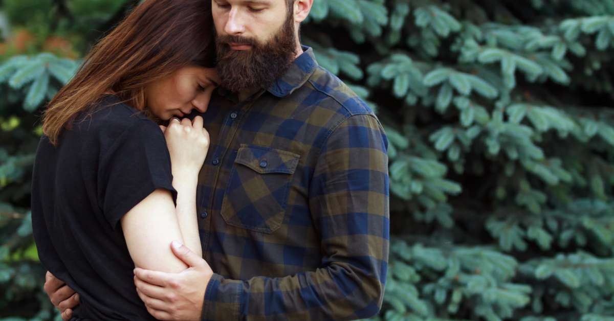 A man hugging a sad woman. They are outside near some plants and the woman is wearing a black shirt and pants.