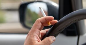 A person holding a lit cigarette inside of a car. Their hand is also on the wheel, and it's a sunny day outside.