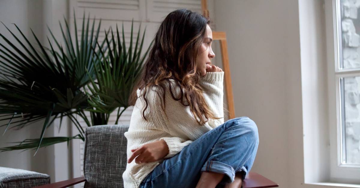 A woman sitting curled up on a chair looking contemplative. There's a plant behind her chair and she's looking out the window.