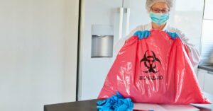 A person wearing a protective suit holds a bag with a biohazard label. There's a fridge in the room.