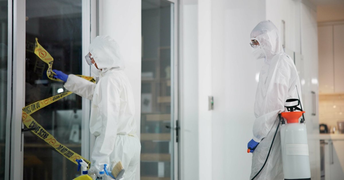 Two people dressed in white protective suits and carrying equipment. One person is removing crime scene tape.