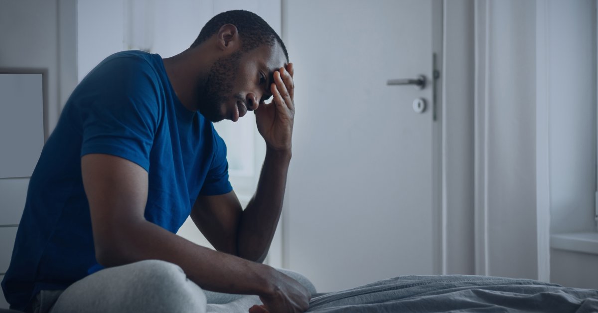 A man sitting on a bed and looking down. He has his hand on his forehead and is wearing a blue shirt.