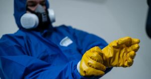 A person wearing yellow gloves, blue personal protective equipment, and a respirator. They are pulling on a glove.