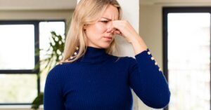 A woman with blond hair holding her nose with her left hand. She's wearing a blue sweater and there's a plant behind her.