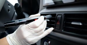 A close-up of a person's hand cleaning the inside of a car with a small brush. The person is wearing a white glove.