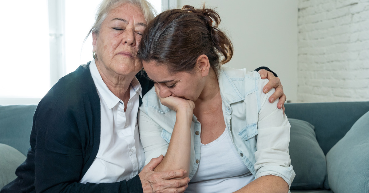 An older woman hugging a younger woman as they sit on a couch. There are pillows on the couch.