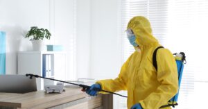 A person in a yellow protective suit and blue gloves sanitizes a wooden desk. There's a bookcase in the background.