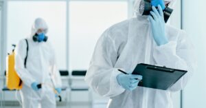 Two men in hazmat gear standing by large windows. One holds a clipboard while the other sanitizes the room.