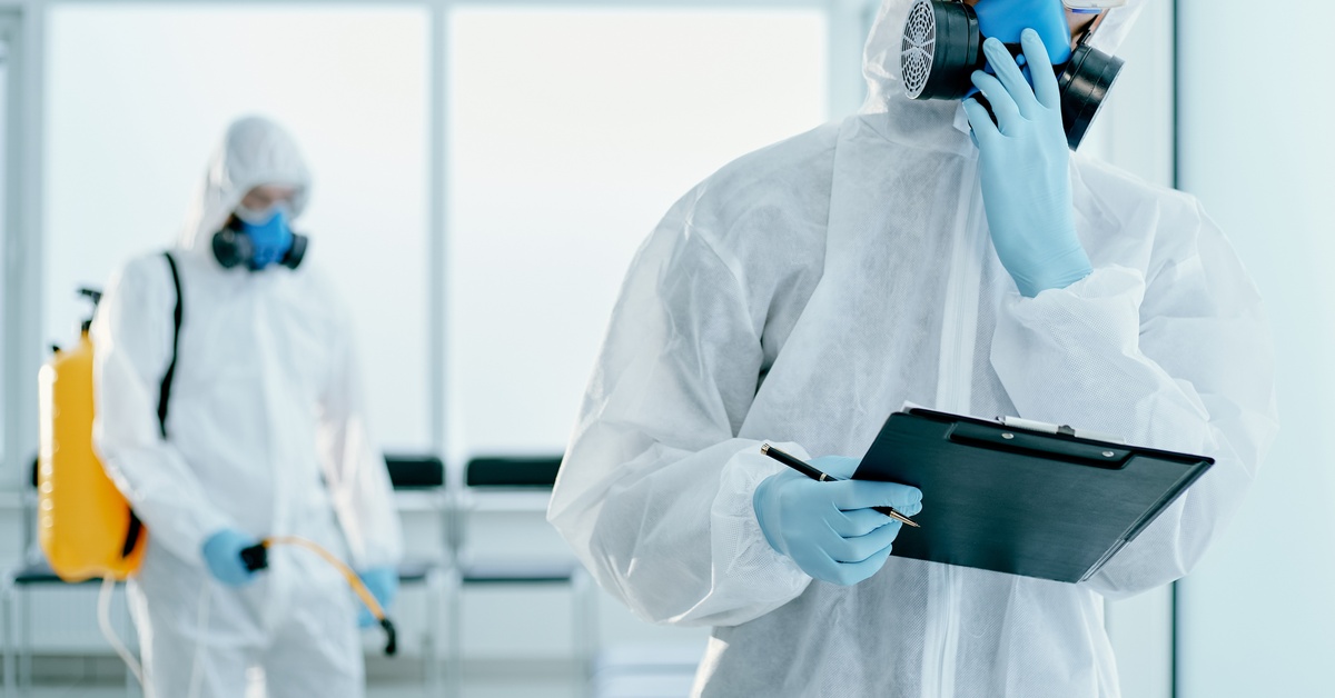 Two men in hazmat gear standing by large windows. One holds a clipboard while the other sanitizes the room.