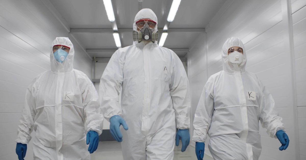 Three hazmat workers walking in a row down a white-walled hallway. The one in the middle wears a gas mask.