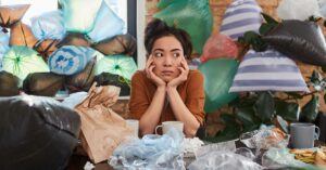 A woman sits with her elbows on a table and her head resting in her hands. She's surrounded by trash and plastic bags.