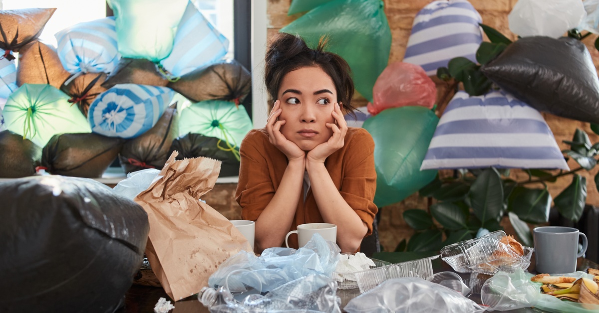 A woman sits with her elbows on a table and her head resting in her hands. She's surrounded by trash and plastic bags.