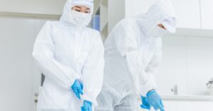 Two people in white hazmat gear and blue gloves standing side-by-side and cleaning a room with white walls and cabinetry.