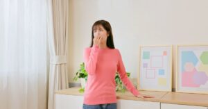 A woman in a pink shirt wrinkling her nose and covering her mouth. She stands near a large wooden dresser.
