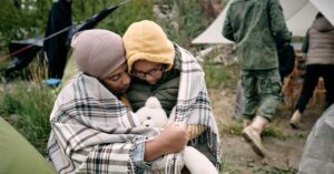 A woman and child crouching and hugging outdoors under a plaid blanket. Behind them, a soldier walks toward a tent.