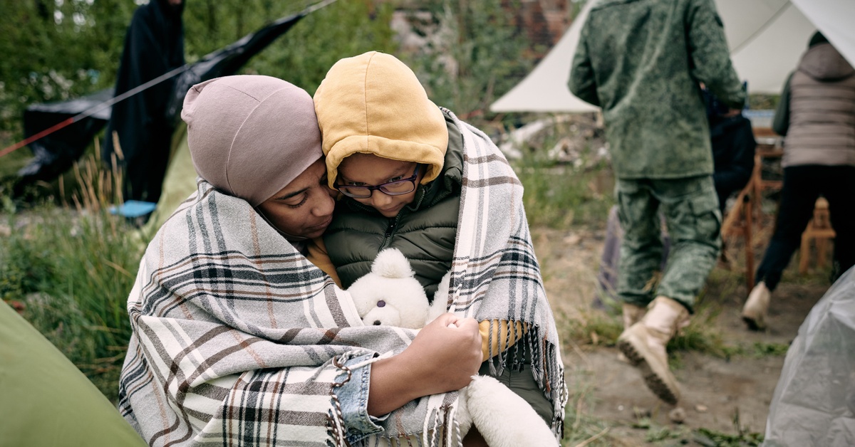 A woman and child crouching and hugging outdoors under a plaid blanket. Behind them, a soldier walks toward a tent.