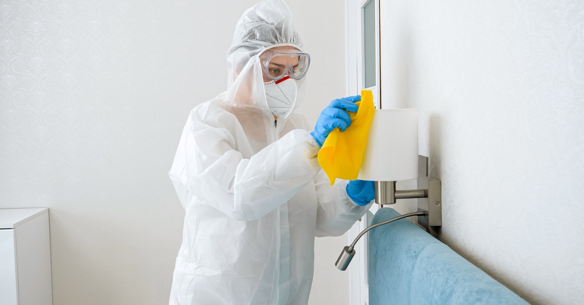 A worker in a hazmat suit, safety goggles, and gloves using a yellow rag to wipe down a lamp installed over a bed.