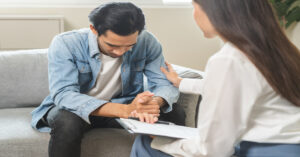 A man sitting on a couch and looking down at his clasped hands while a woman with a clipboard touches his arm supportively.
