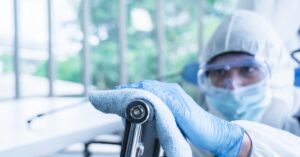 A person in a hazmat suit, safety goggles, and gloves using a blue rag to wipe the stainless steel back of a chair.