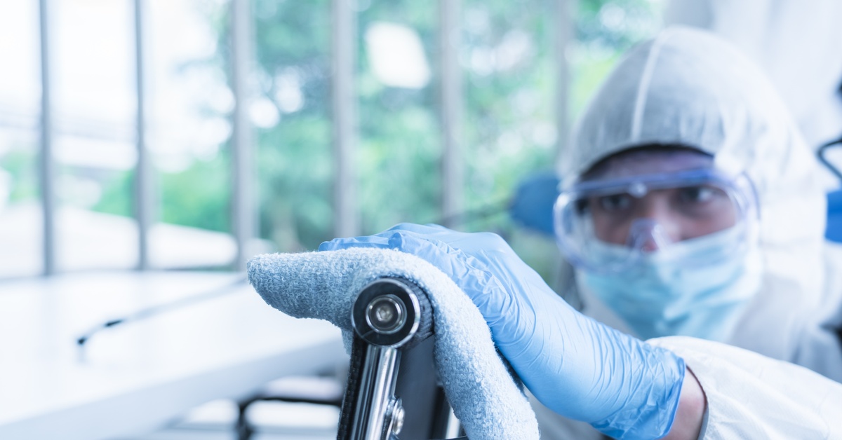 A person in a hazmat suit, safety goggles, and gloves using a blue rag to wipe the stainless steel back of a chair.