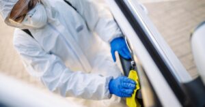 A hazmat worker in a protective suit and gloves using a yellow rag to clean the interior of a white car.