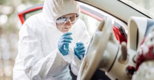 A woman in a white hazmat suit, safety goggles, and gloves collecting samples from the interior of a red car.