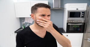 A bearded man in a black T-shirt stands inside a kitchen with one hand covering his mouth and nose.