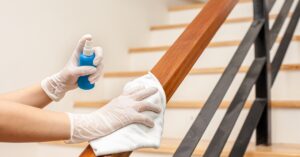 White-gloved hands using a small blue spray bottle and a thick white rag to clean a wooden stair railing.