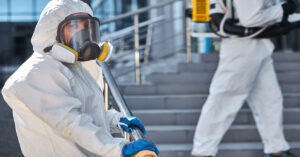 A worker in a white hazmat suit holding a steel stair railing outdoors. Another suited worker stands further up the steps.