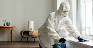 A worker in a white protective suit and black gloves using a rag and blue cleaning fluid to disinfect a windowsill.