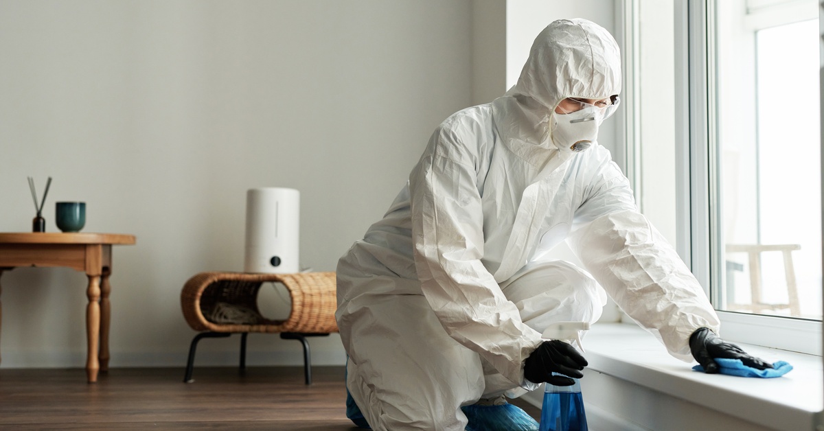 A worker in a white protective suit and black gloves using a rag and blue cleaning fluid to disinfect a windowsill.
