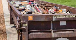 A brown, large dumpster parked in a driveway and filled to the brim with trash and discarded items.