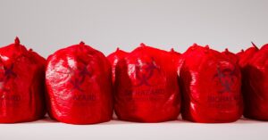 A line of red, disposable biohazard waste bags, filled and tied off at the top against a white wall and floor.