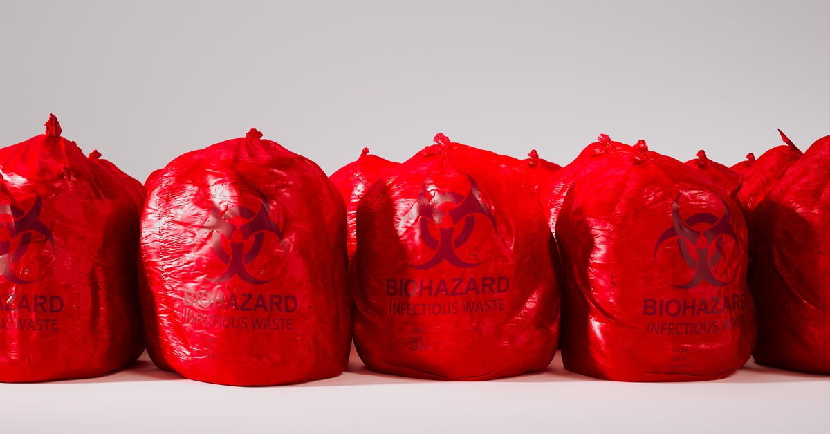 A line of red, disposable biohazard waste bags, filled and tied off at the top against a white wall and floor.
