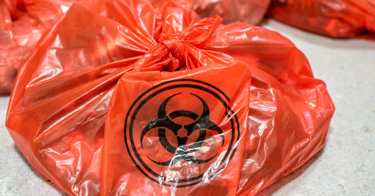 A red hazardous waste bag on the floor next to a pile of identical bags in the background. Its contents are obscured.