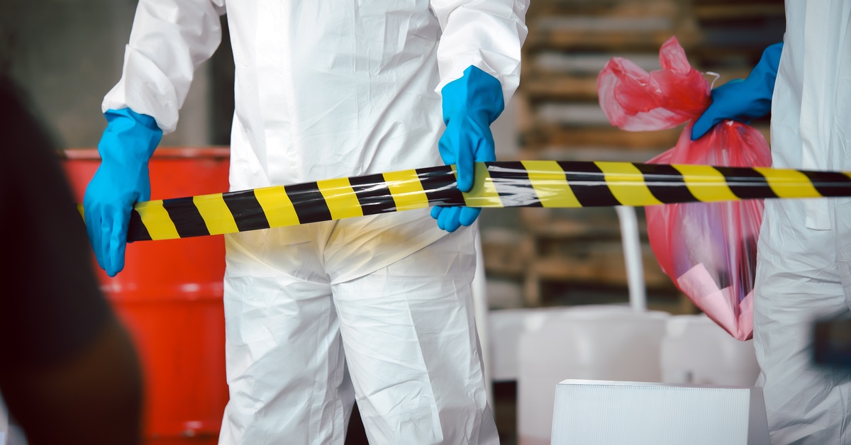 Two people in protective suits at a work site. One person holds a red trash bag and the other pulls out caution tape.