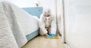 A cleaning person wearing a full-body suit, mask, and gloves, wiping the floor of a bedroom with a yellow rag.