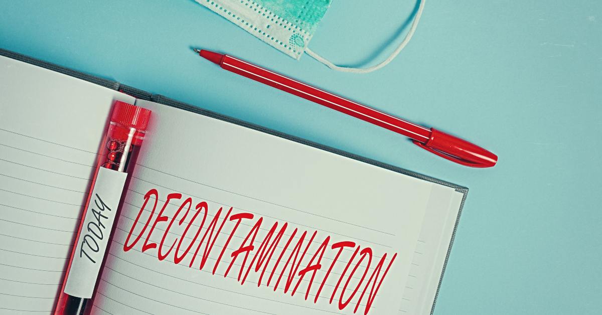 A close-up of a notebook labeled "decontamination" in all capital letters with a red pen, mask, and blood vial next to it.