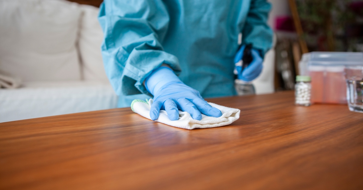 A close-up of a person wearing a blue full-body protective suit and gloves, wiping the surface of a table with a cloth.