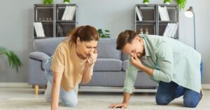 A man and a woman kneeling on a carpet in their living room, holding their noses and making displeased faces.