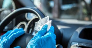 A close-up of gloved hands cleaning a steering wheel with a disinfecting wipe inside a car's cabin.