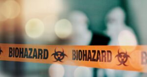 A close-up of a biohazard orange tape stretched across an opening with blurry shapes of people in the background.