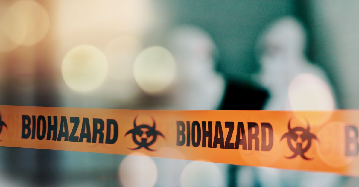 A close-up of a biohazard orange tape stretched across an opening with blurry shapes of people in the background.